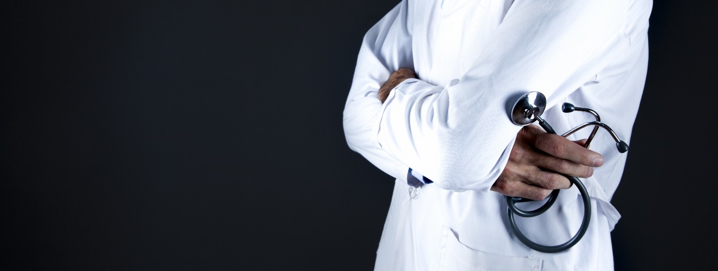 A soctor in a traditional white jacket, arms crossed and a curled stethoscope in his/her right hand. We see the clinician from waist to upper chest level. There is a black background.