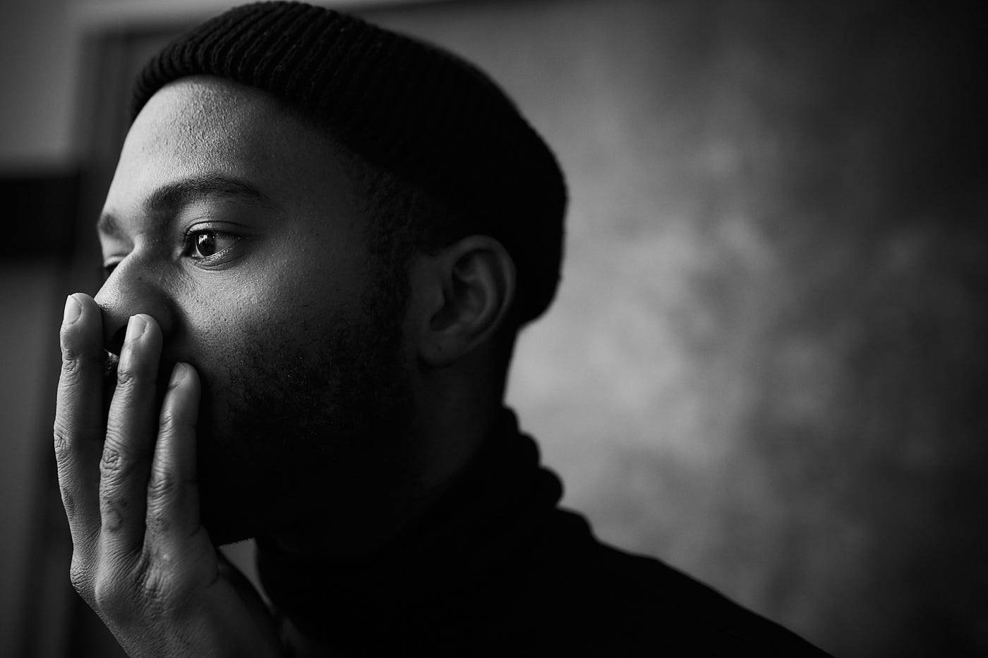 As seen in profile to the left of the image, a black man in his 30s faces left, his right hand extended up to cover his mouth. The image is in black and white.