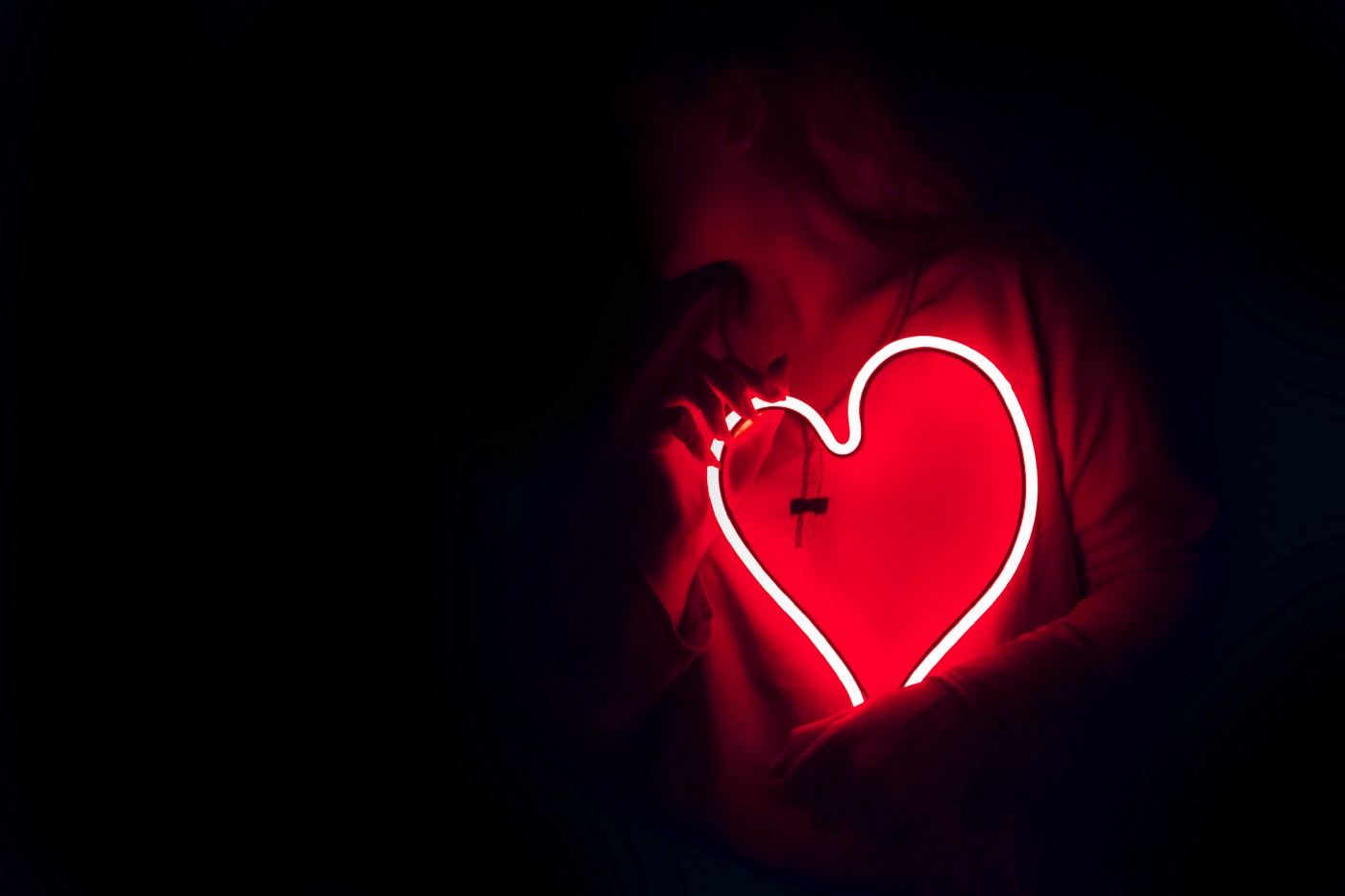 Woman, on the right side of the screen, holds a heart-shaped fiberoptic line over her torso. Black background, with woman barely in view.