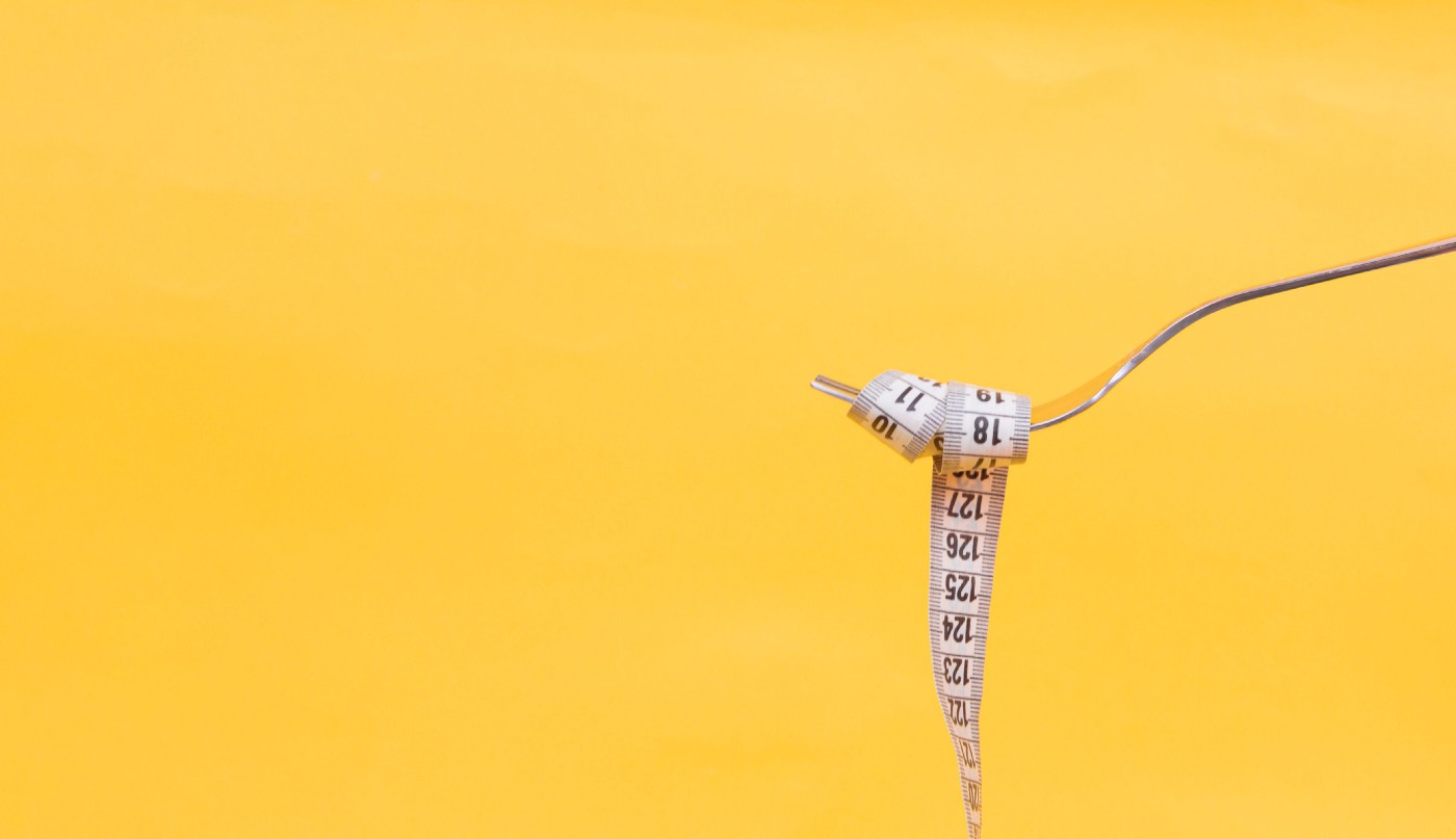 Fork, extending from the right of the picture, holds a measuring tape, looped around the tines and extending to the bottom of the image. Yellow background.