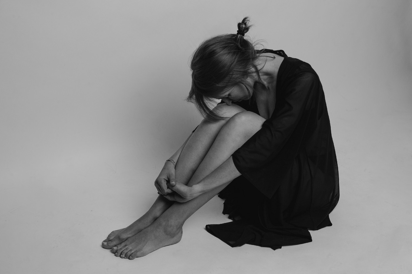 Black and white image of a young woman sitting on the floor, leaning forward to clutch her knees.