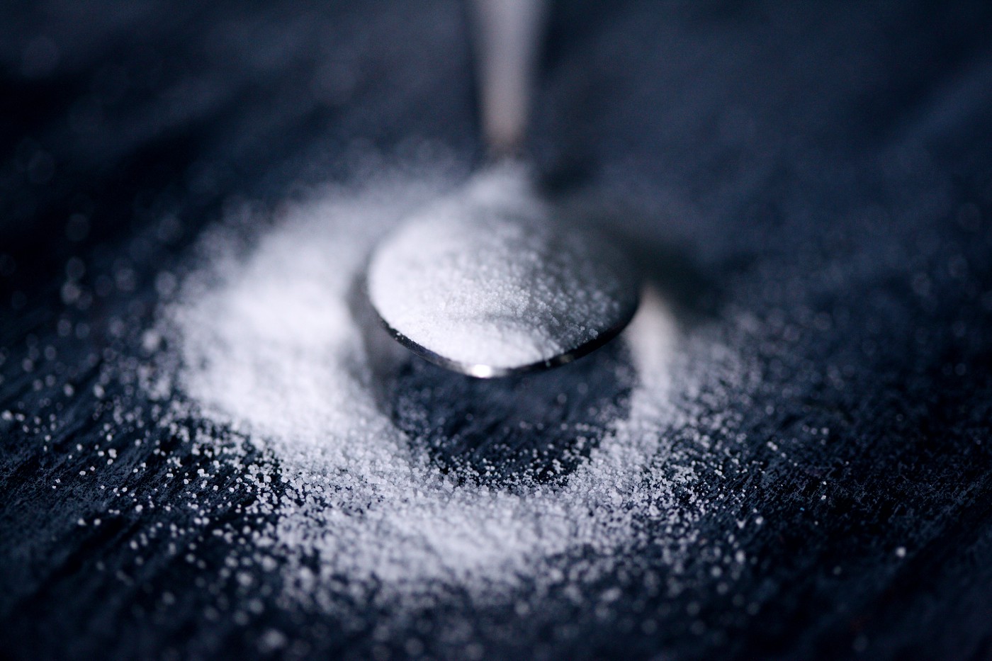 A photo of a spoon with some sugar. Beneath the spoon, on the black tabletop, is more sugar. Eating more refined carbohydrates (or other things with lots of sugar) is associated with higher triglyceride levels.