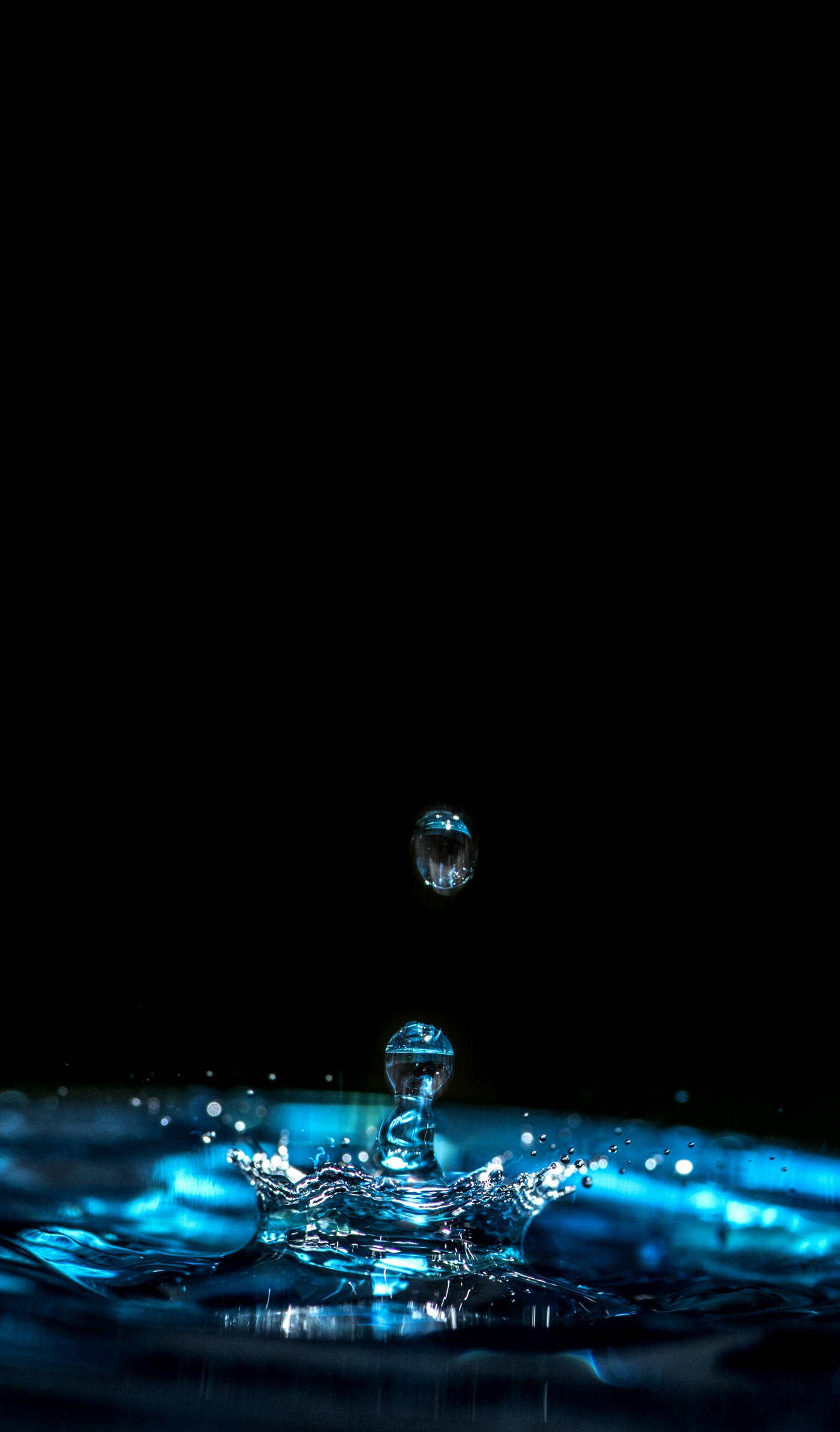 A photograph of a drop of water hitting a pool of water and bouncing up off of it. Black background.