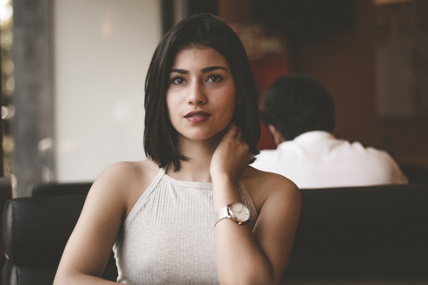 Young brunette woman, in her 30s, looks directly at the camera. A man is seated to the right in the distance, facing away. The woman wears a sleeveless white top.