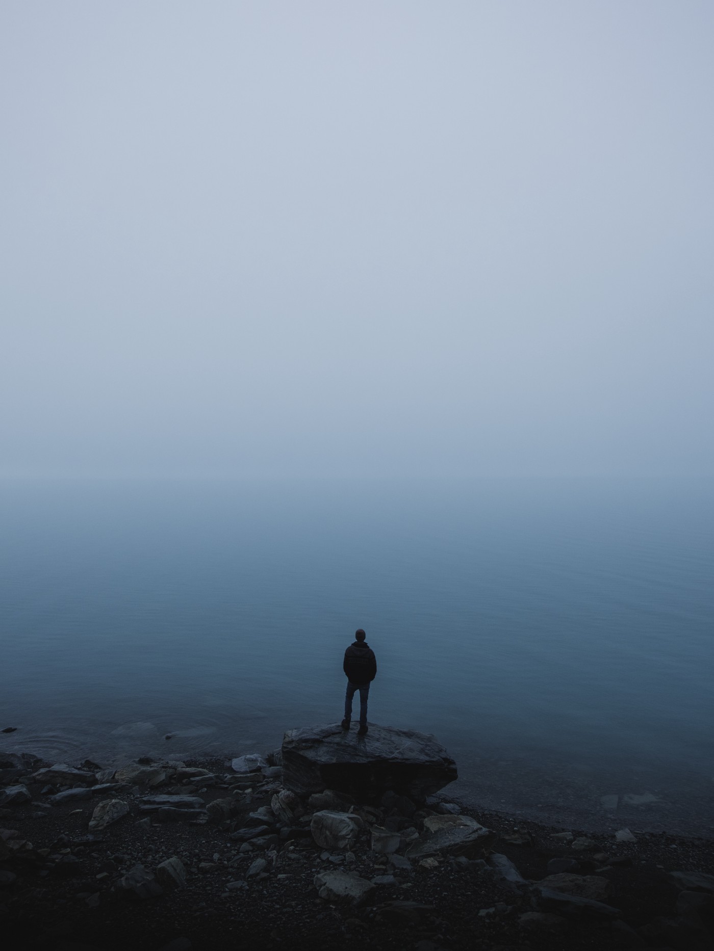 A man stands on a rock, looking at the ocean in the distance. The scene is a hazy blue.