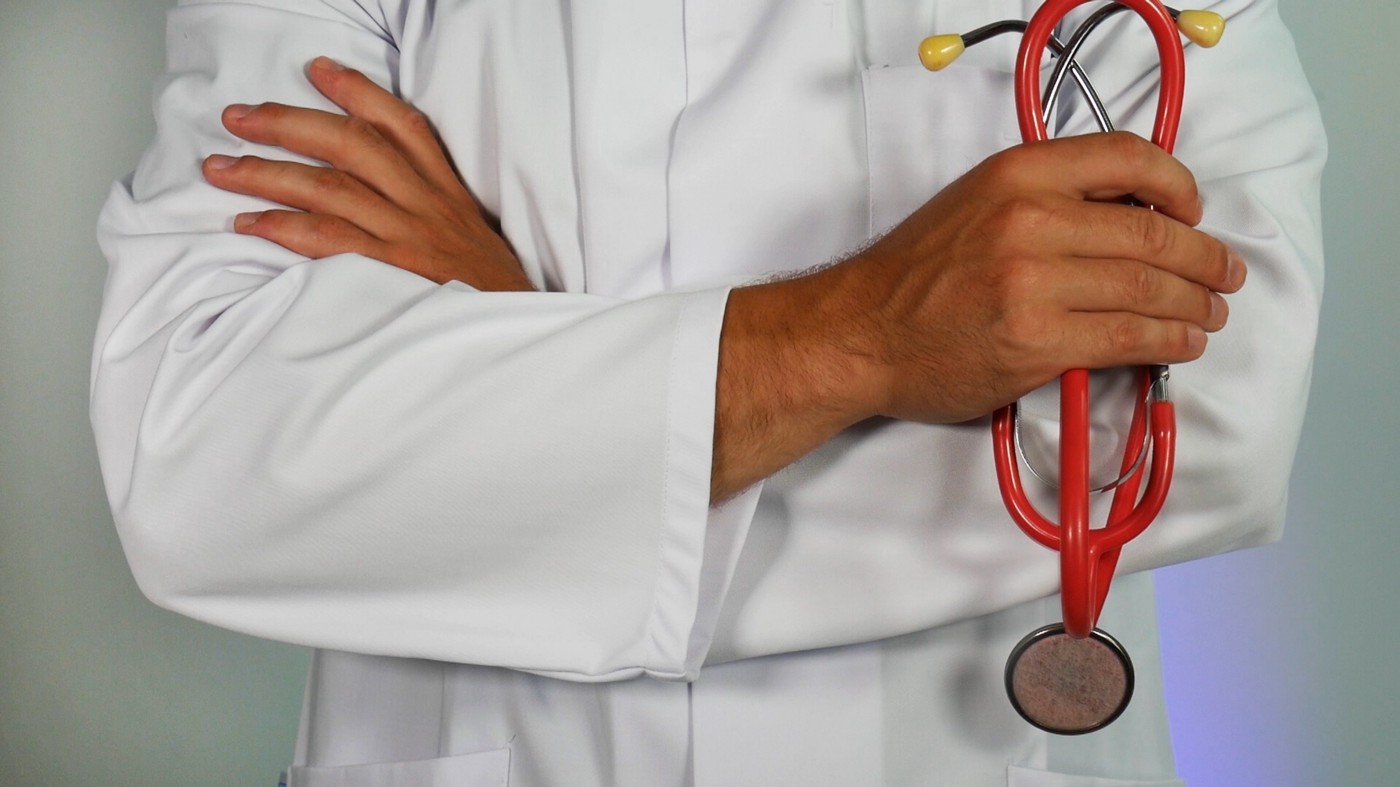 White coat-clad doctor faces us, hands across his chest. In his right hand, he holds a stethoscope. We see only his mid torso.