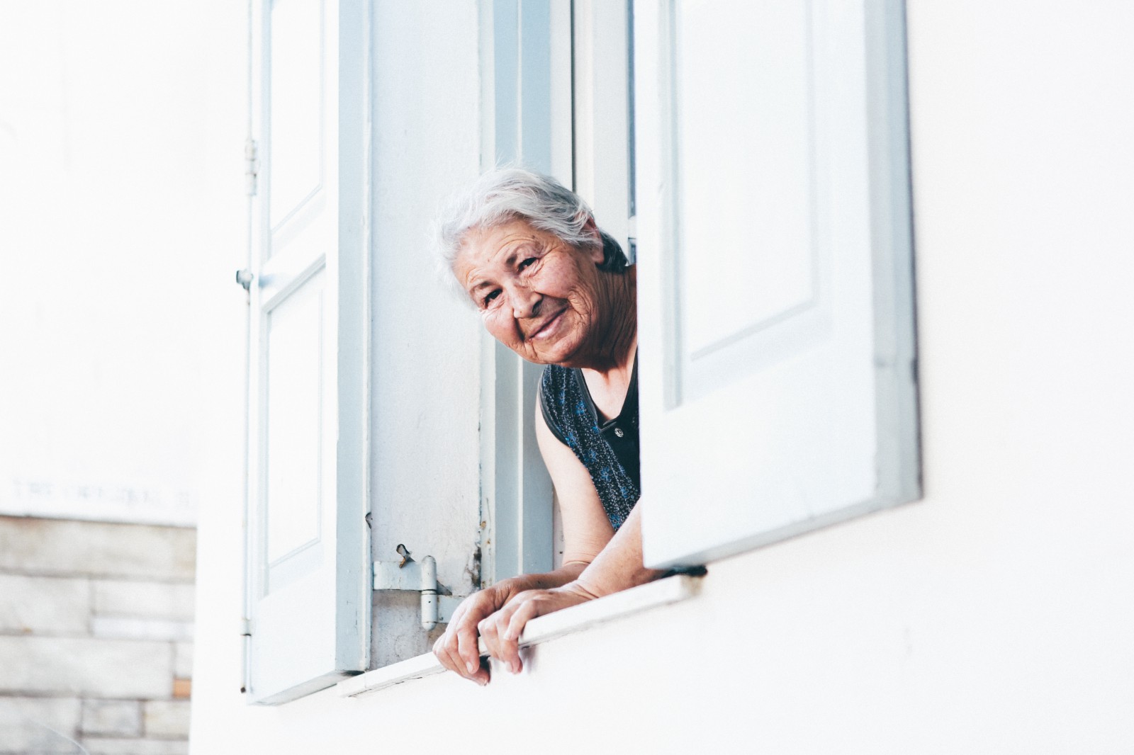 An older woman leans out of a window of a white house. She is percehed between two white shutters.