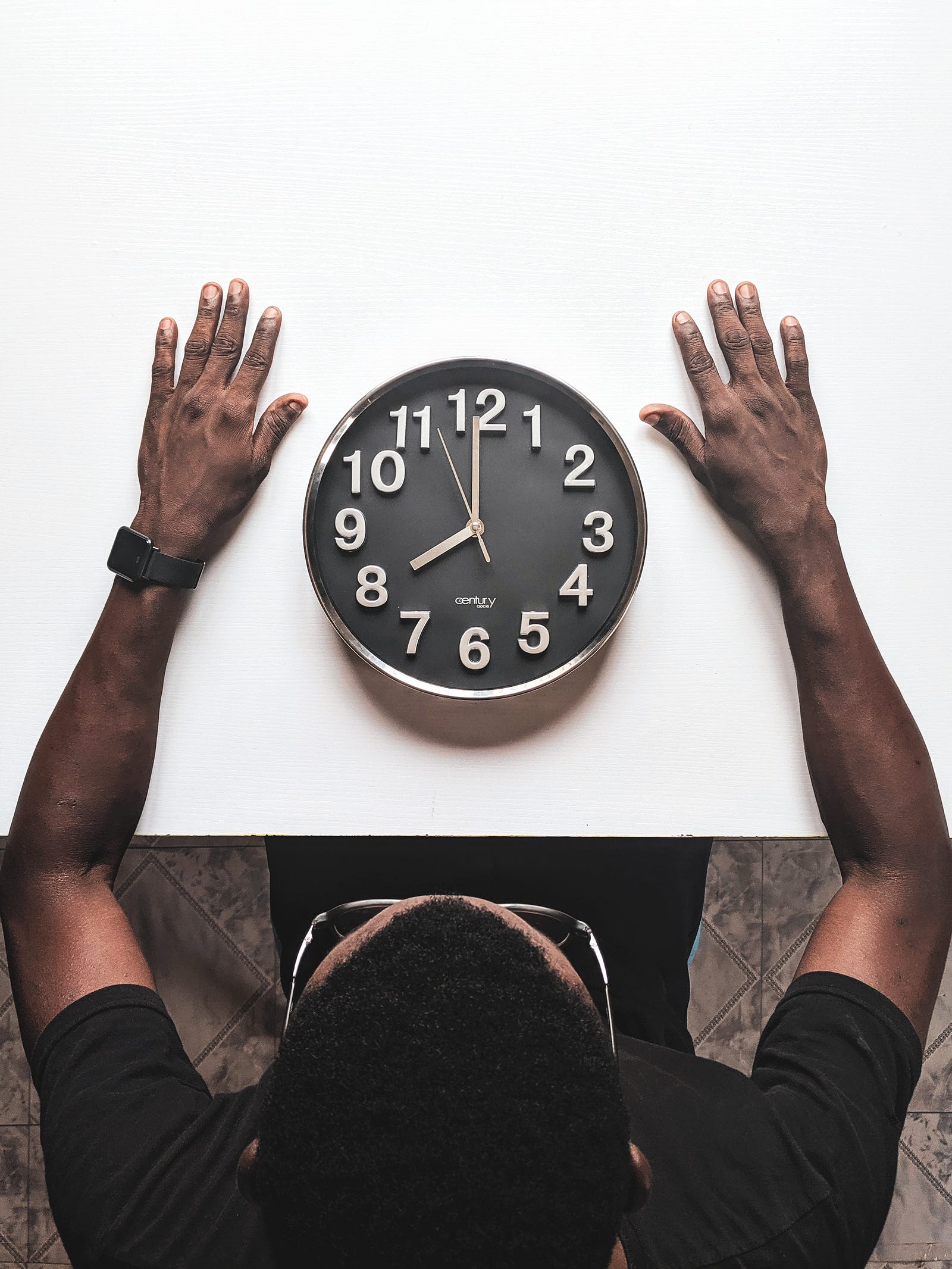 A clock with a black face *and white numbers and hands) sits on a white table. A person extends their arms around the clock. Why might vaccination effectiveness be linked to the time of day? Researchers think that the circadian rhythm — 24-hour cycles that are part of the body’s internal clock, running in the background to carry out essential functions and processes — affects the body’s response to the vaccine.
