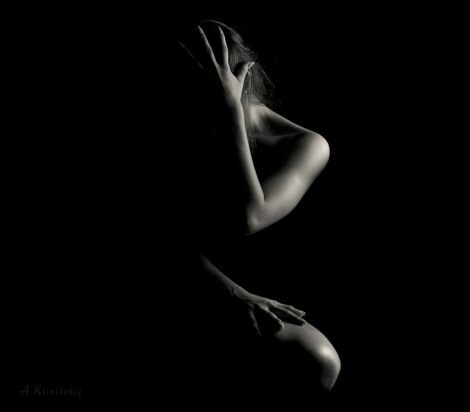 A nude woman faces us with her right arm covering her face . The image is dark, but we see her left side highlighted by light.