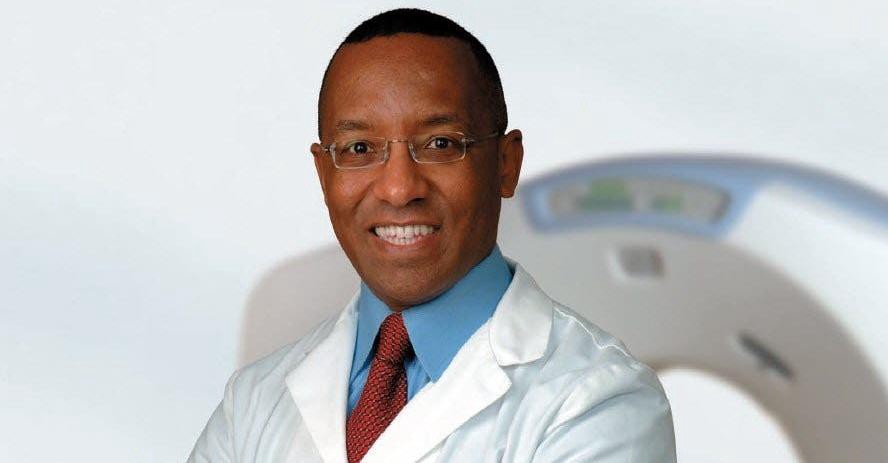 Dr. Michael Hunter dons a white medical jacket. He wears a blue shirt, red tie, and rimless eyeglasses. In the background, we see a white CT scanner.