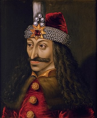 Painting of Vlad the Impaler. He wears a brown curly wig that extends below his shoulders and a hat with a star on the front. Vlad has a long moustache.