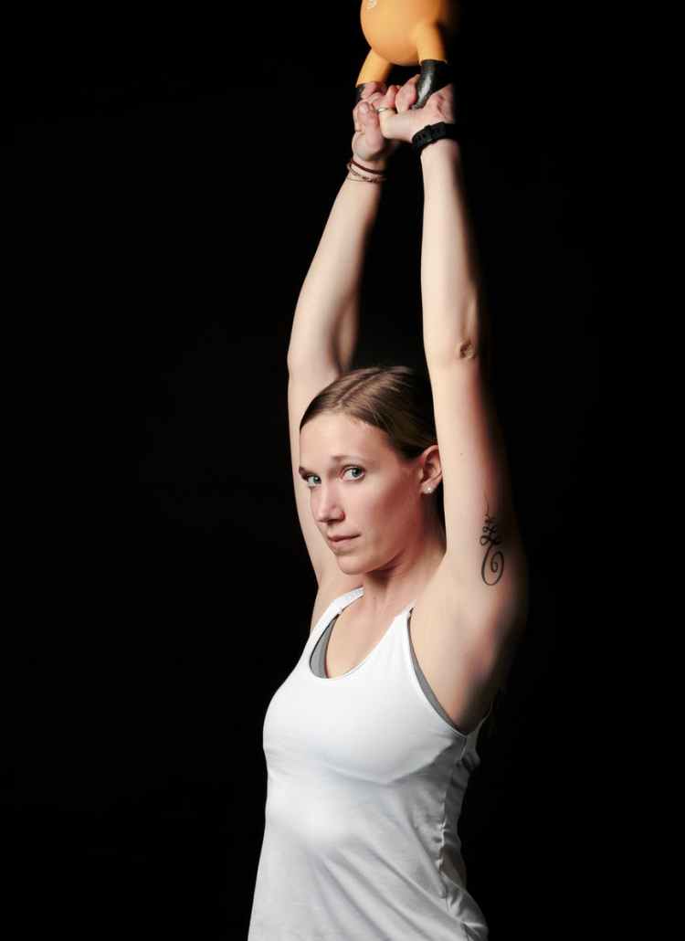 side view photo of woman in white tank top lifting orange kettle bell standing against black background