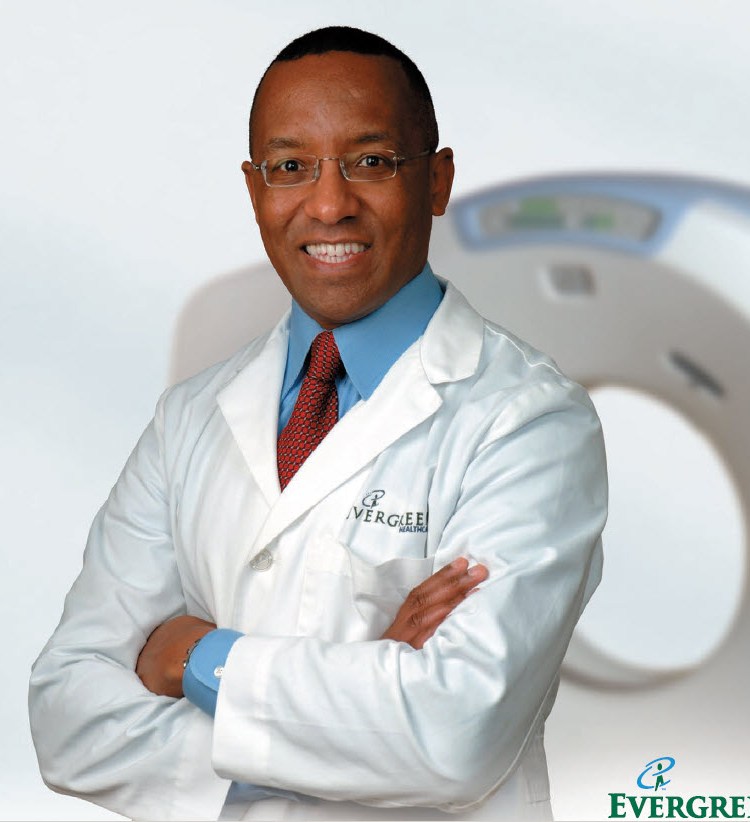 Portrait of Dr. Michael Hunter, MD, wearing a white medical coat and smiling with arms crossed in front of a CT scanner.