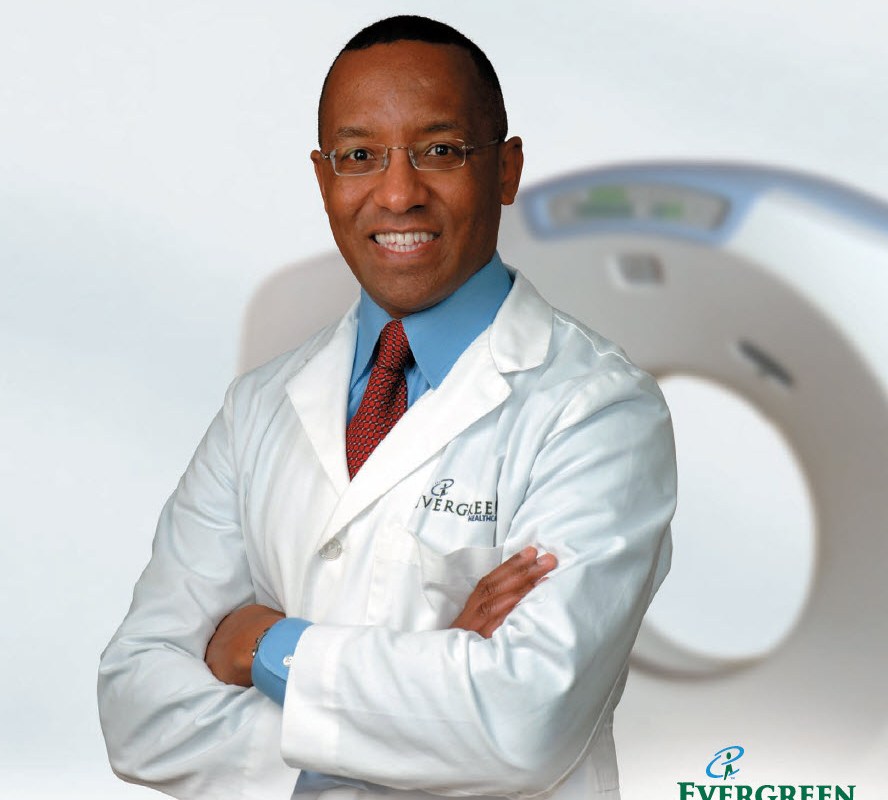Portrait of Dr. Michael Hunter, MD, wearing a white medical coat and smiling with arms crossed in front of a CT scanner.