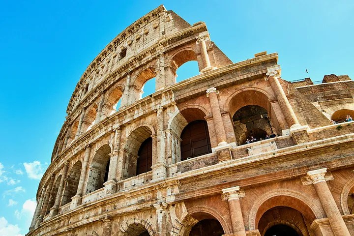 Low-angle view of the Colosseum in Rome under a bright blue sky, showcasing its ancient arches and weathered stone.