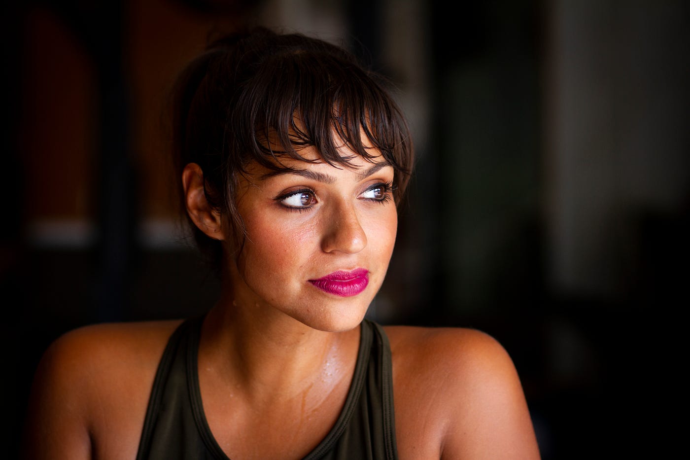 A young brunette woman looks off to her left as she faces us. She wears a tank top and we see her from upper chest up. Dark background. A recent systematic review and meta-analysis discovered that strength training could reduce blood pressure.